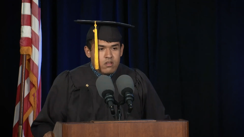 A student from Riverview in his graduation cap and gown, speaking at the podium.