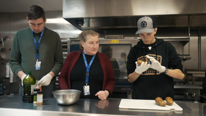 Two students work on culinary skills with a staff member in school kitchens.