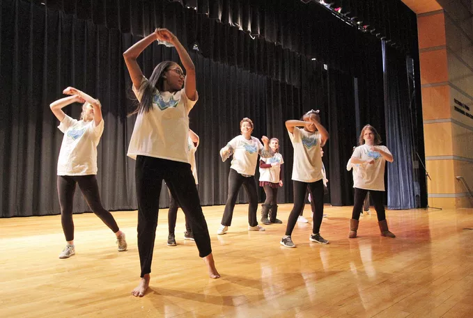 Several students in yellow t-shirts and black pants dance on a stage.