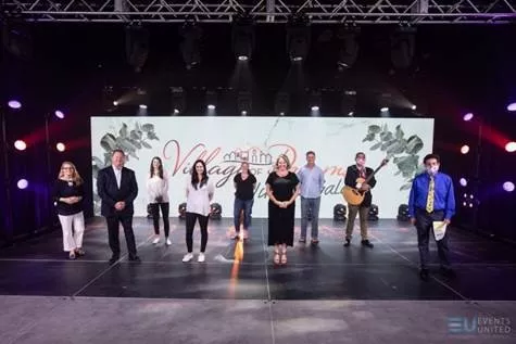 Nine people standing in a studio stage with a screen in the bacground that reads "Village of Dreams Virtual Gala".