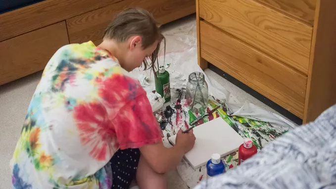 A student in the process of painting on a canvas on the floor of her dorm.